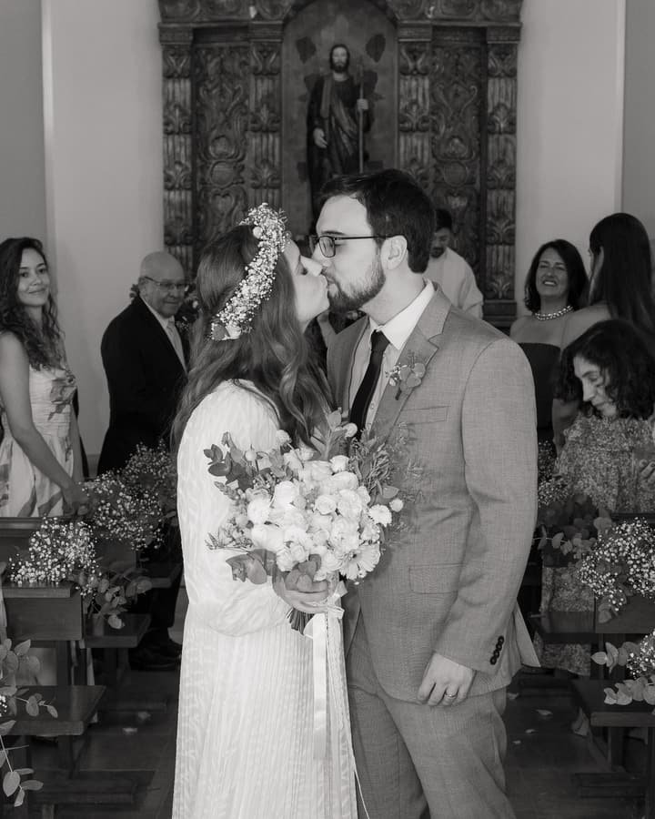 Beijo dos noivos no altar, em preto e branco, com os convidados ao fundo